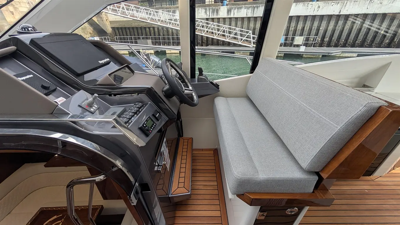Modern boat cockpit featuring a steering wheel, control panel, and a grey cushioned seat against a backdrop of a marina.